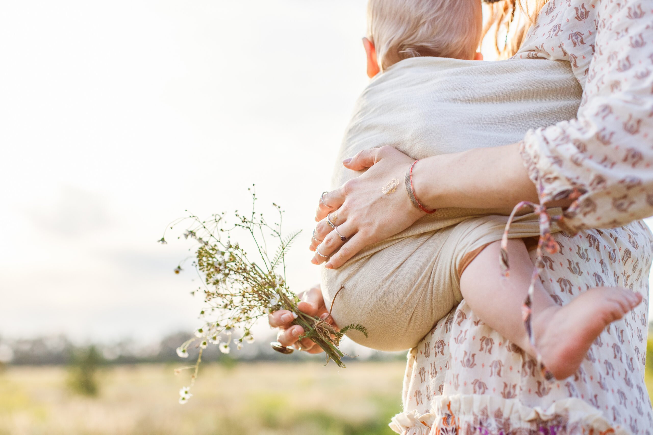 Little baby boy and his mother walking in the fields during summer day. Mother is holding and tickling her baby, babywearing in sling. Natural parenting concept