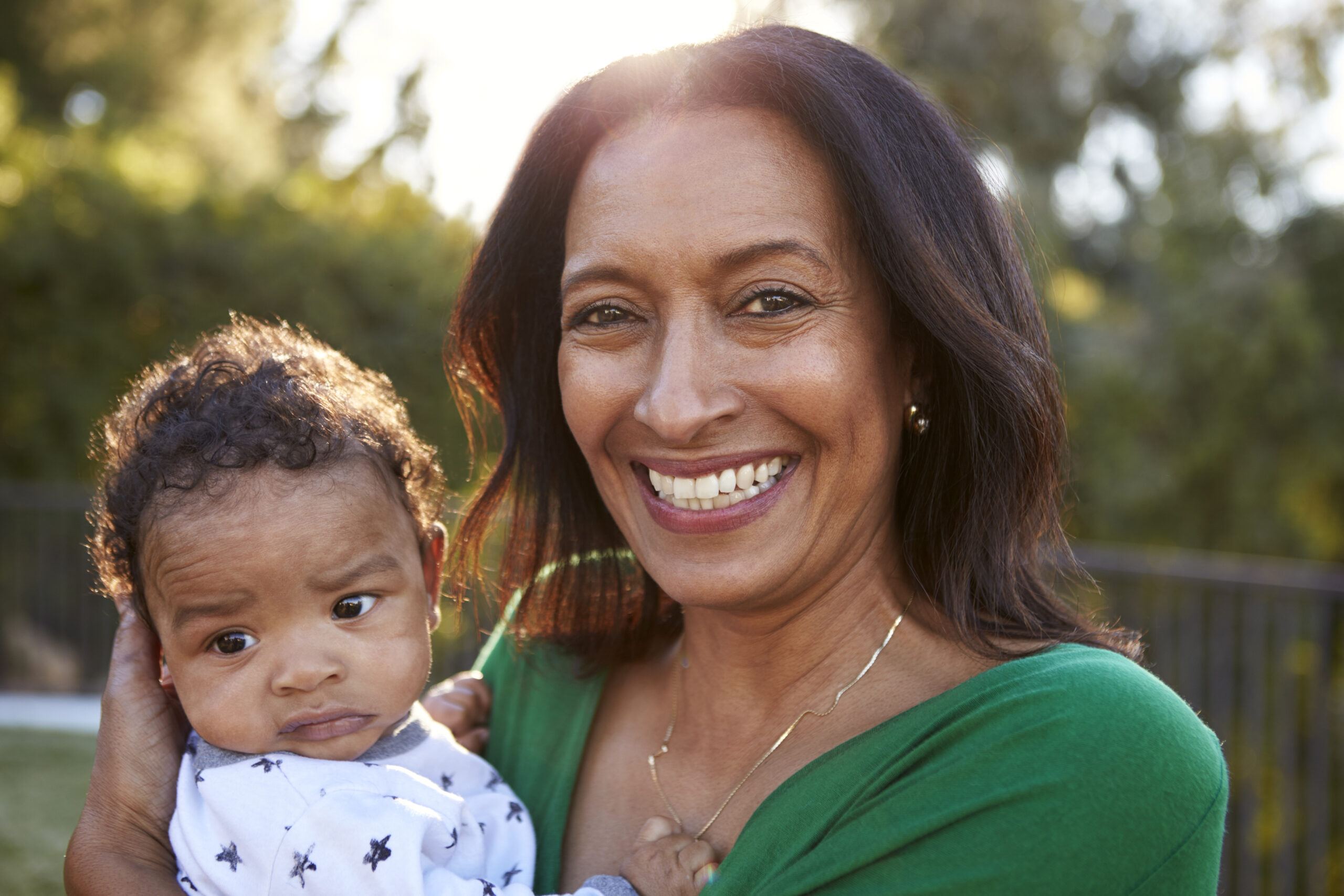 Happy mixed race middle aged grandmother holding her three month old grandson in the garden and smiling to camera, close up, head and shoulders