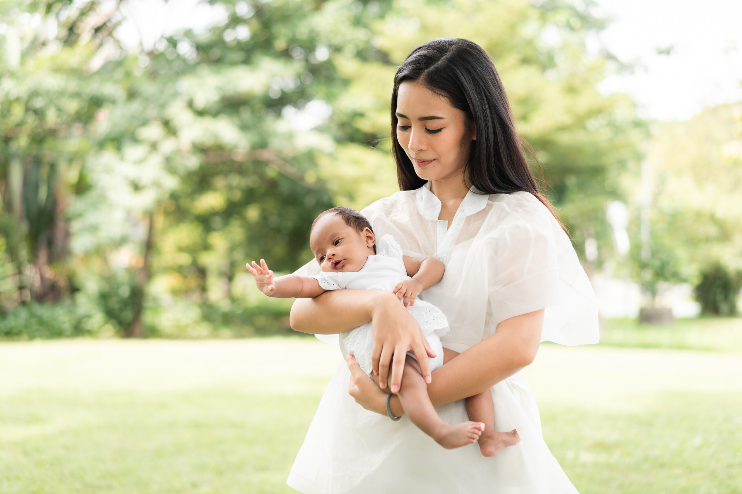 Asian young beautiful mother holding her newborn is sleeping and feel with love and touching gently then sitting on green grass in the park