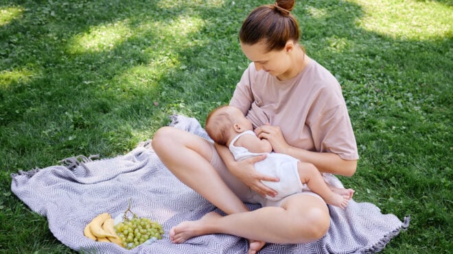 Cheerful mother breastfeeding her newborn son during family picnic in the park sitting on blanket surrounded by green grass on summer day with her child looking satisfied